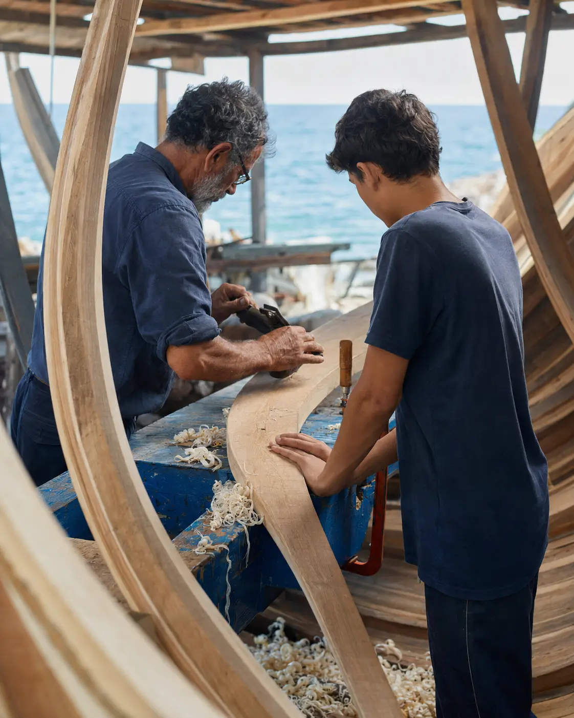 Boatbuilder and apprentice shaping a curved timber plank inside a coastal yard